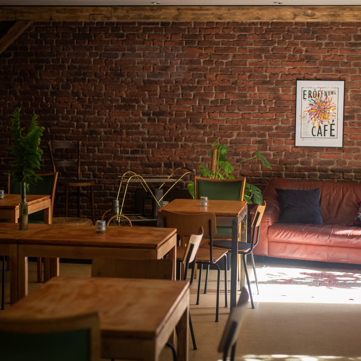 Photo showing a cafe scene with three tables, two wooden, one green chair, and a sofa in the back right. In the left corner is a small stage before a red brick wall.